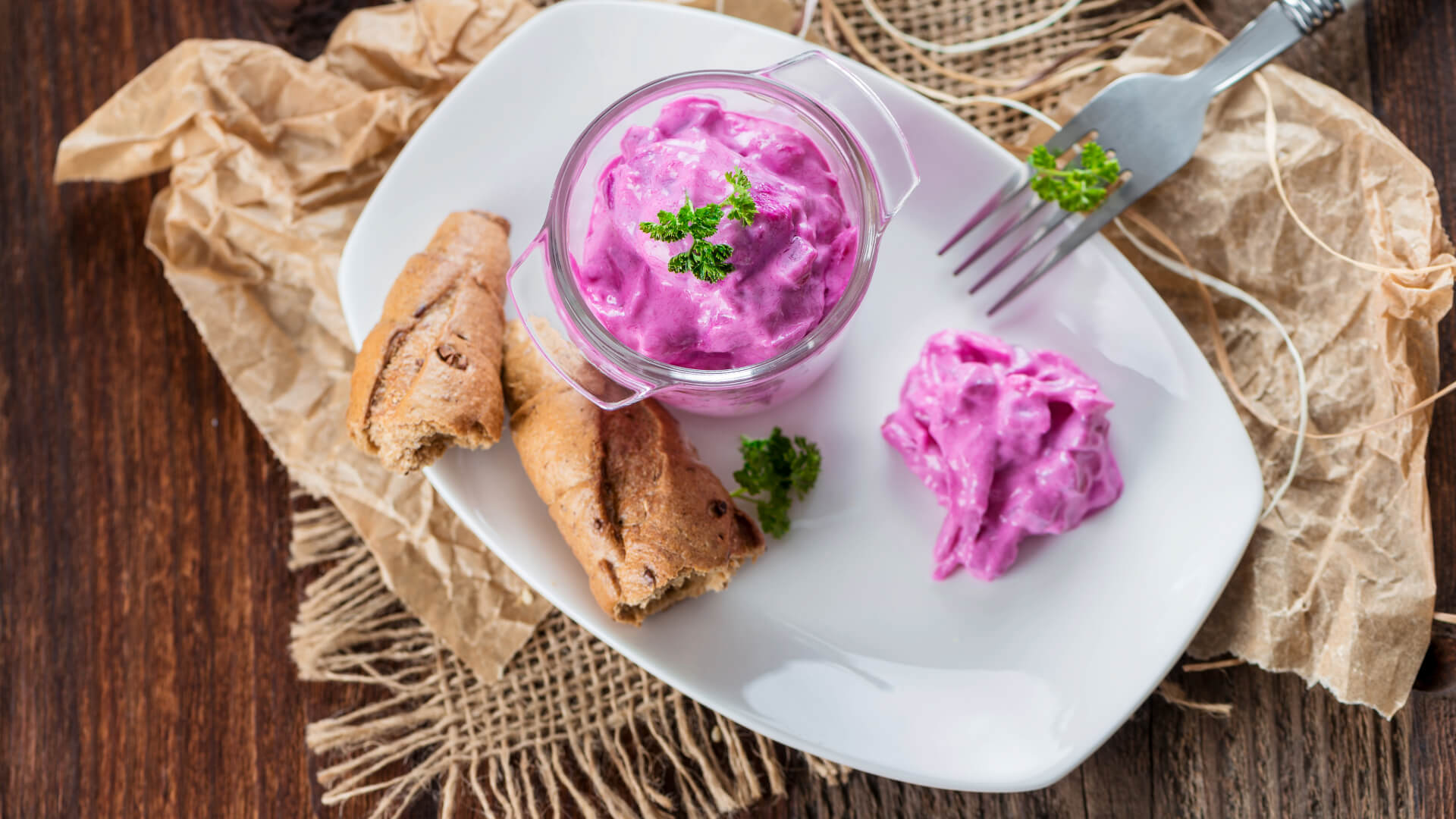 in Glas mit cremigem Heringssalat in kräftigem Pink, garniert mit frischer Petersilie, serviert auf einem weißen Teller mit knusprigem Brot und einer kleinen Portion Salat daneben. Rustikale Tischdekoration mit Jutestoff und Pergamentpapier im Hintergrund.